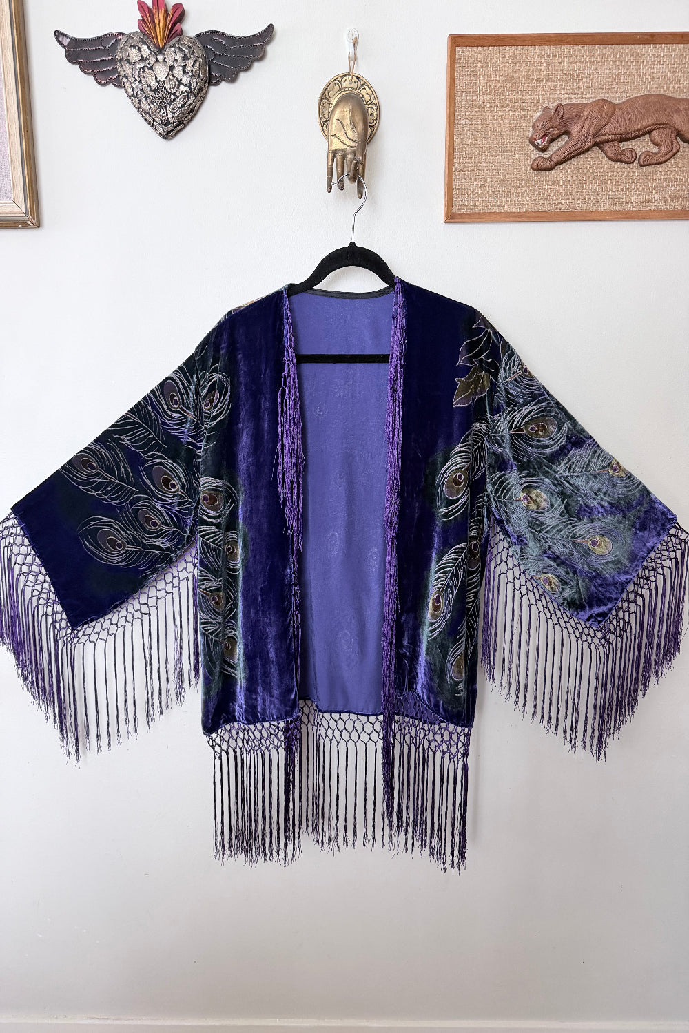 Peacock feather print purple velvet silk jacket with fringe on a hanger against a white wall.
